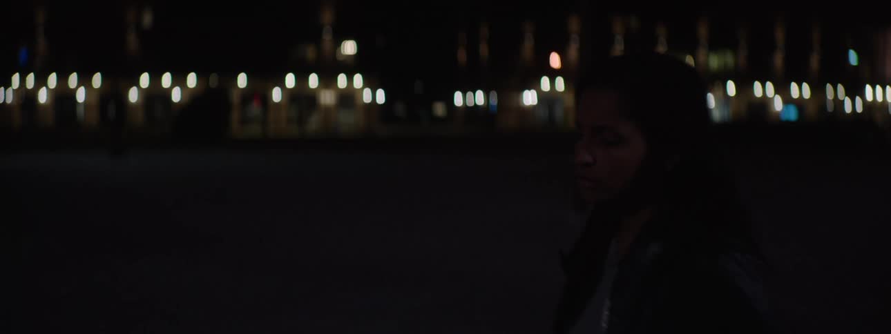 In the video a woman with long hair is walking on a city street at night she approaches a silver car opens the drivers side door and gets in
