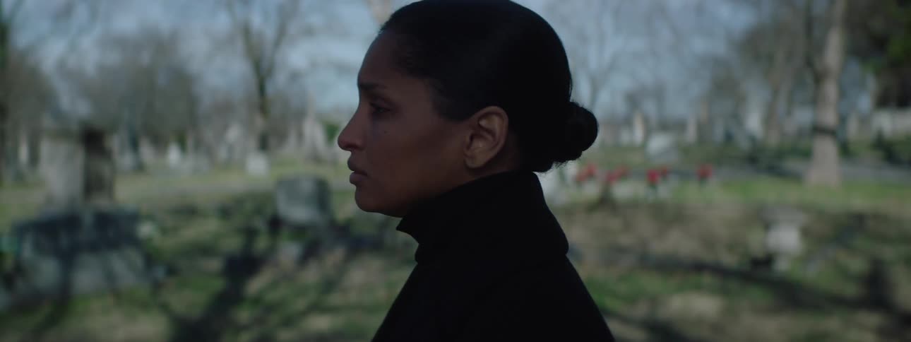 Young woman dressed in all black standing in a cemetery and watching a funeral service from a distance filled with sadness and shedding tears