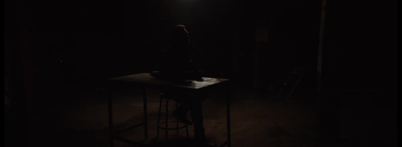 In a dark room with dramatic lighting a person is seen sitting on a stool at a table