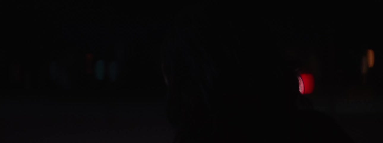 A close side view of a womans face is shown in a dark city street with blurred orange and white lights in the background