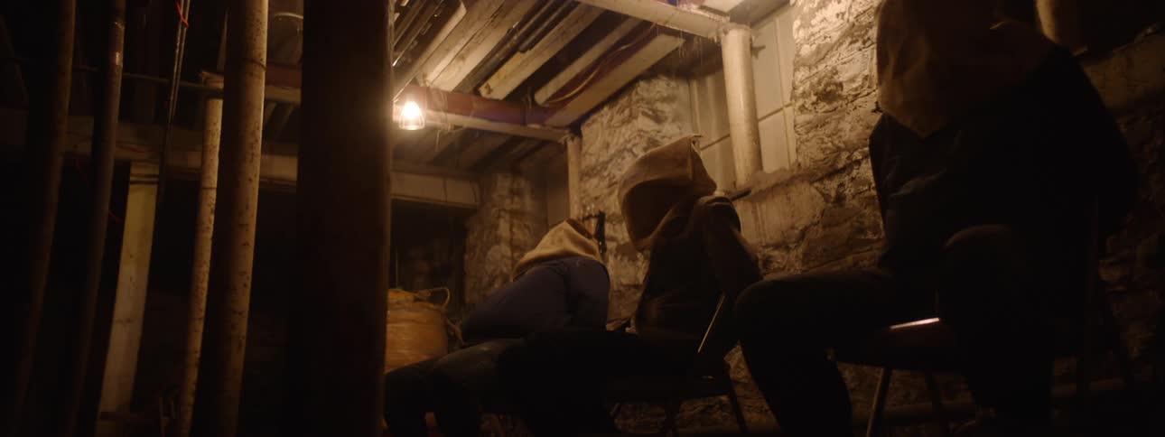 In the video the screen is initially dark before revealing a dimly lit dungeonlike room with concrete walls several people are sitting in chairs with what appear to be hoods or bags covering their heads
