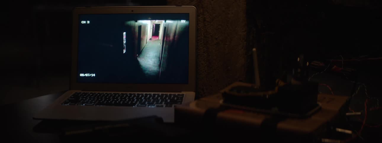 In a dark room a laptop displays a live security camera feed of a long empty hallway creating a tense and suspenseful surveillance scene