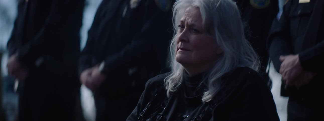 Elderly woman sitting in a cemetery at midday during a funeral service looking sad with friends and family standing behind her