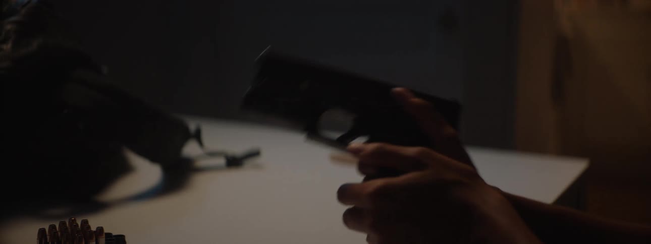 A persons hands are loading and cocking a gun in a dark room before setting the gun on a table