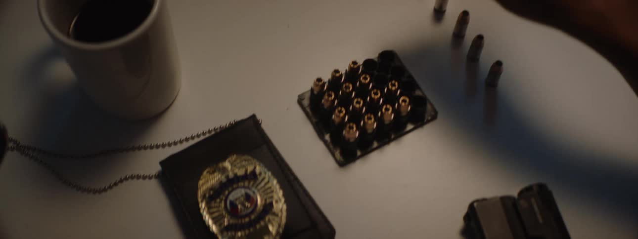 A persons hand is taking a bullet from a tray of ammunition on a table next to a police badge and a handgun