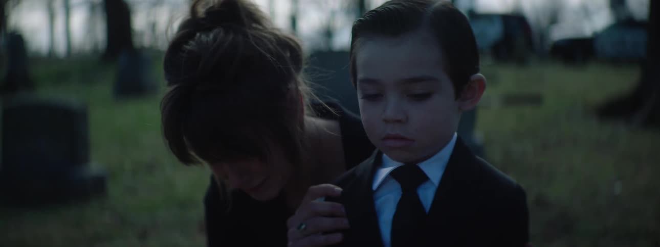 A sad woman wearing all black is holding a young boy in a black suit and white shirt with a black tie as they stand in a grassy cemetery next to a funeral display with flowers and a portrait
