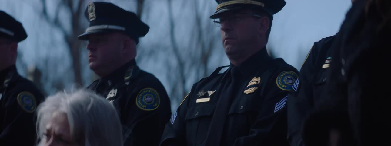 A line of police officers in dark uniforms and caps stands outdoors in a serious atmosphere