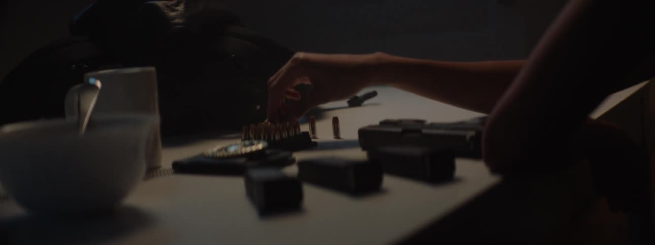 In the video a persons hands are shown loading a magazine with bullets on a table next to the magazine a white bowl and mug are visible with the scene lit by a soft warm light