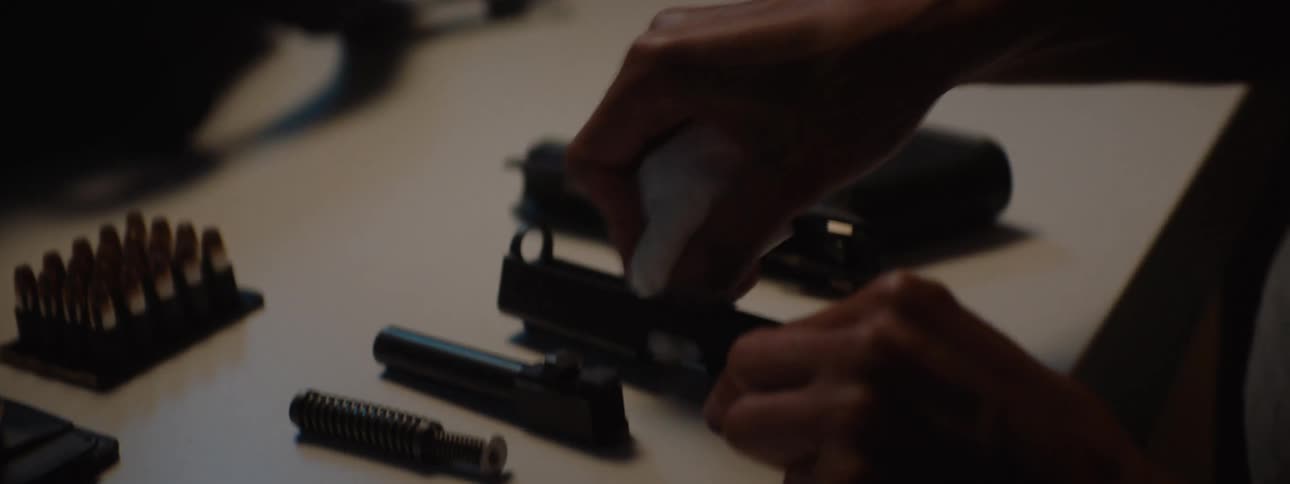 Hands cleaning disassembled gun parts next to ammunition and a badge
