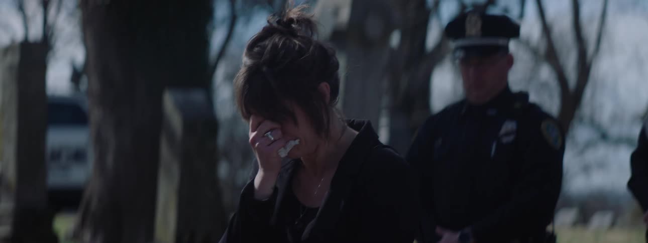 In the video a woman with her hair in a messy bun is seen looking distressed and wiping tears from her face she is standing outdoors with a police officer blurred in the background suggesting a somber or difficult moment