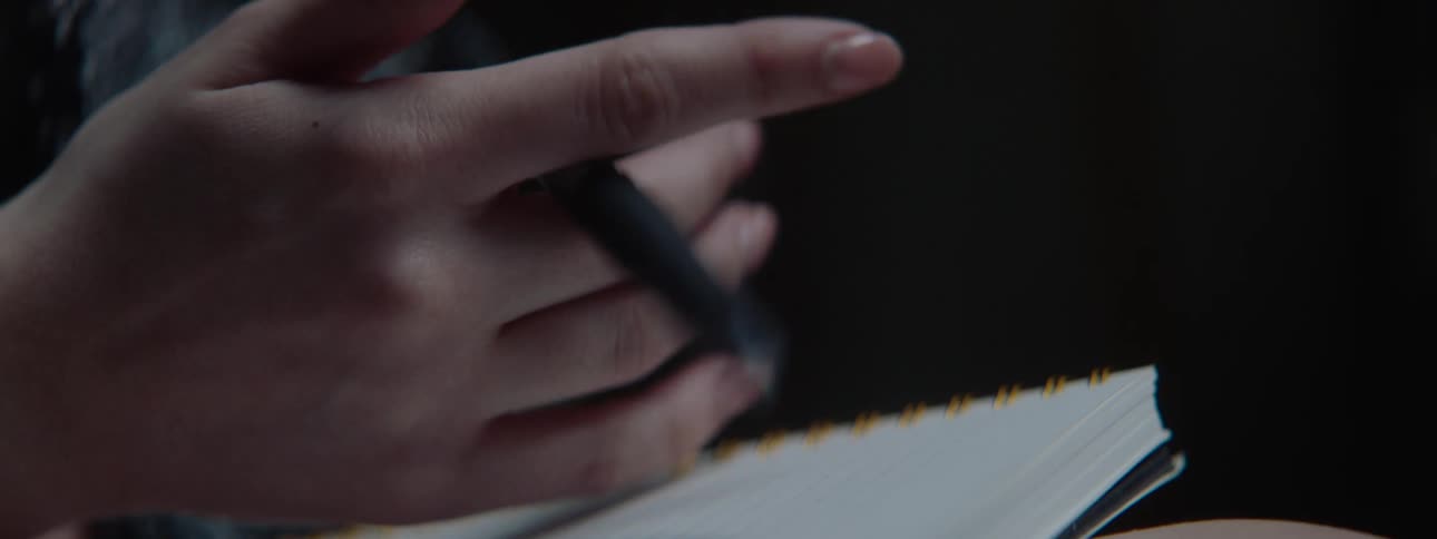 A close view shows a persons hands balancing a black pen over a white spiral notebook with a dark background