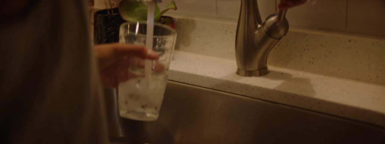 A young black woman picks up a glass cup and goes to rinse it with water in the kitchen sink at night