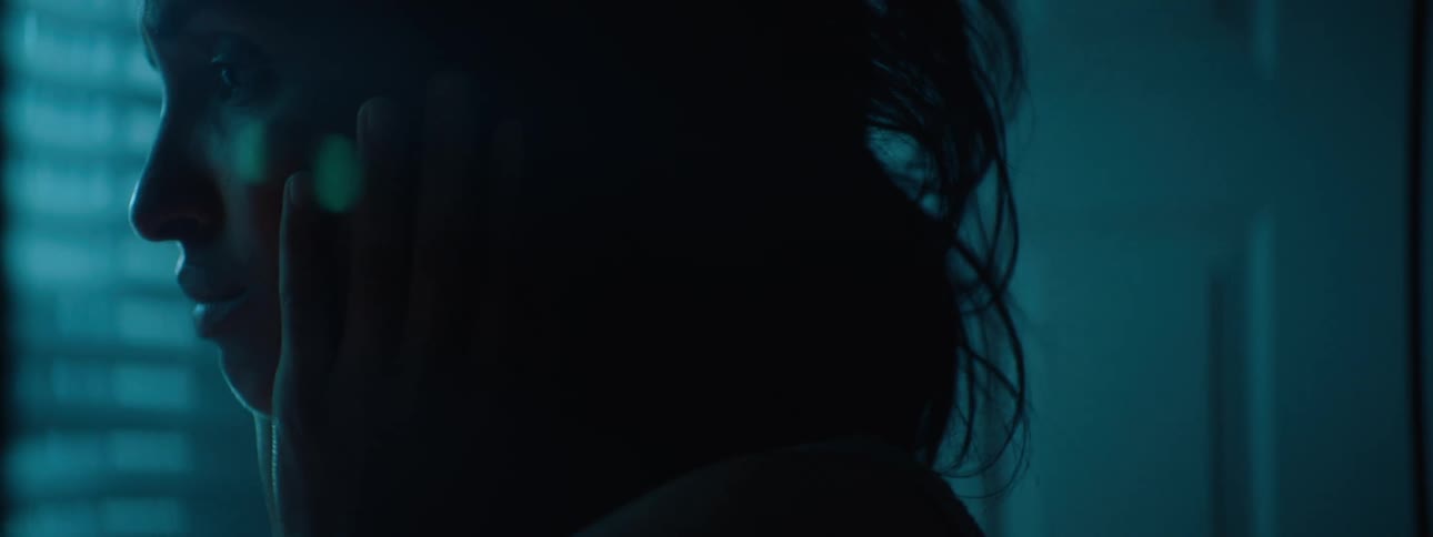 A sad woman with loose hair gazes pensively toward window blinds while touching her face her profile bathed in cool blue light