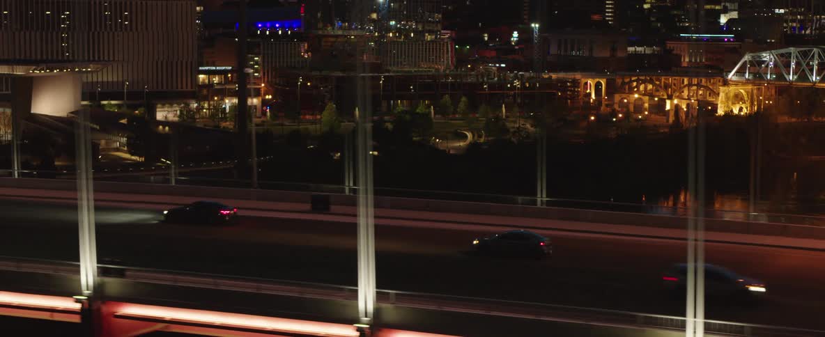 A camera panning from a road with passing vehicles to reveal a city skyline with lights reflecting on the water