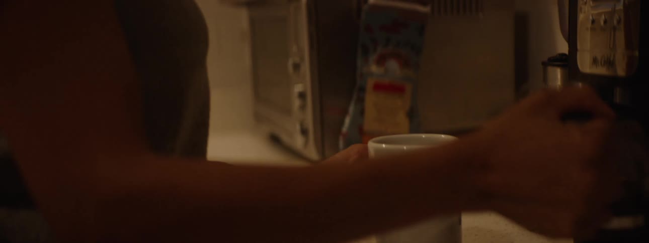 A person pours coffee into a white mug takes a sip walks with a black bag places the bag in a trash can and then walks with a dish toward a table with a vase of flowers