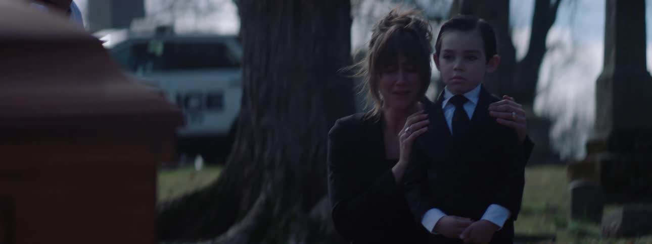 A woman stands with a boy places a hand on his shoulder and wipes tears from her face in a graveyard setting