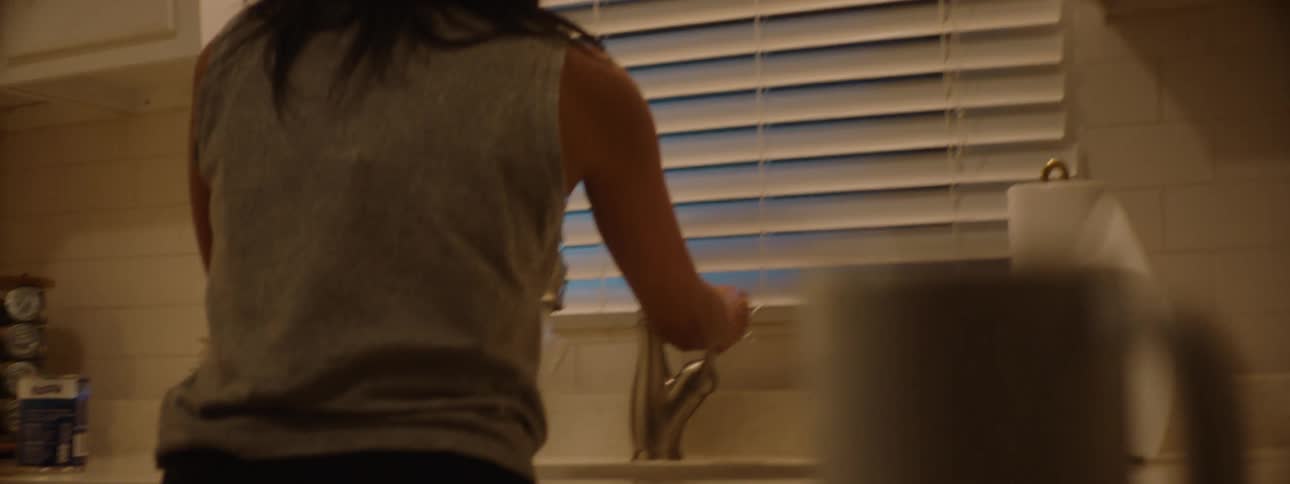 A young black woman picks up a glass cup and rinses it with water at the sink in the kitchen