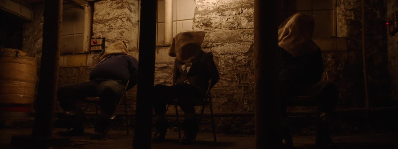 A group of people with sacks over their heads are sitting in a dimly lit dark room with brick walls
