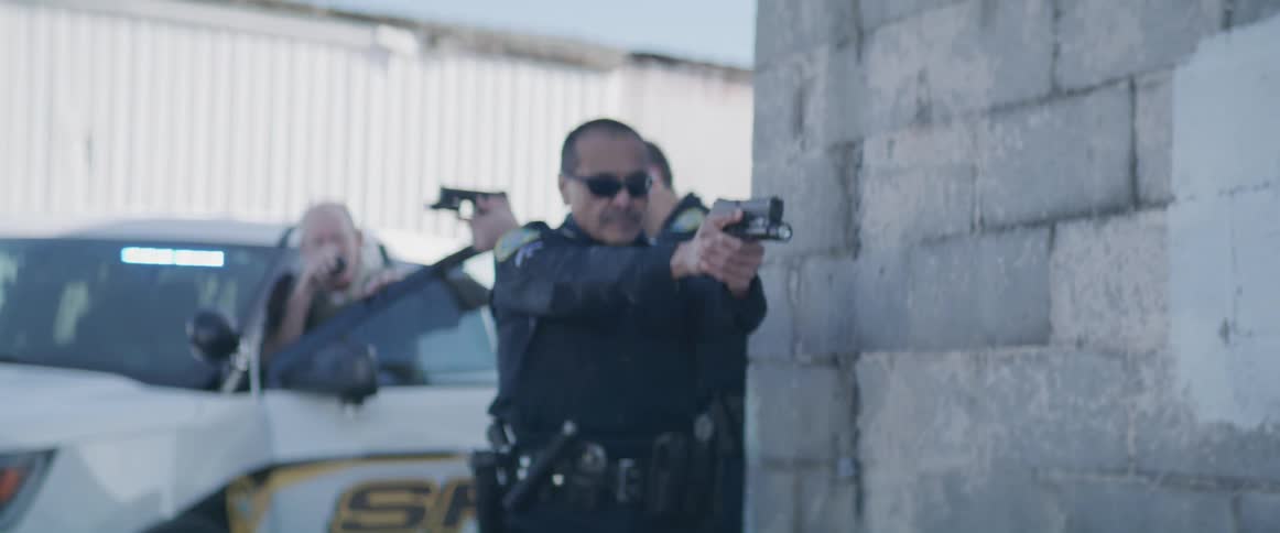 Two male police officers in uniforms and sunglasses take cover behind a white sheriffs car they both aim their pistols at an unseen target
