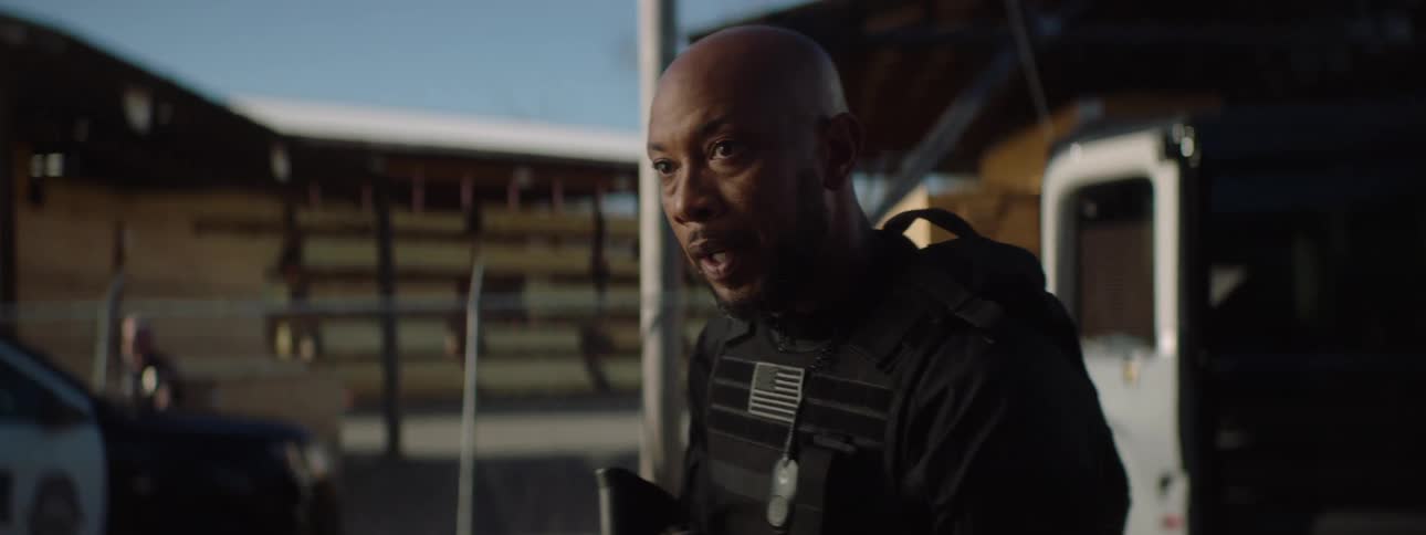 In the video a bald man in a black tactical vest with an american flag patch is shown outdoors he looks serious and is speaking with other armed officers visible in the background behind him