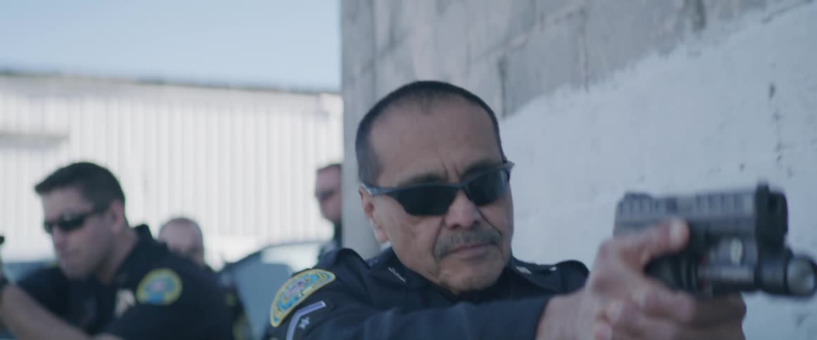 In the video a police officer with a mustache and sunglasses is standing and holding a gun he is aiming it at something offcamera with other police officers visible in the background