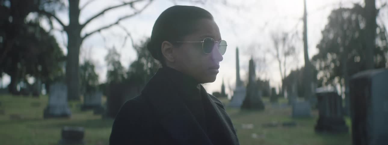 Woman wearing sunglasses walks alone among tombstones in a cemetery