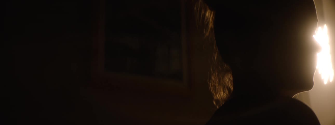 A close side view shows the silhouette of a person in a dark room with long hair as a bright light shines from the side and a glowing object is visible in the background