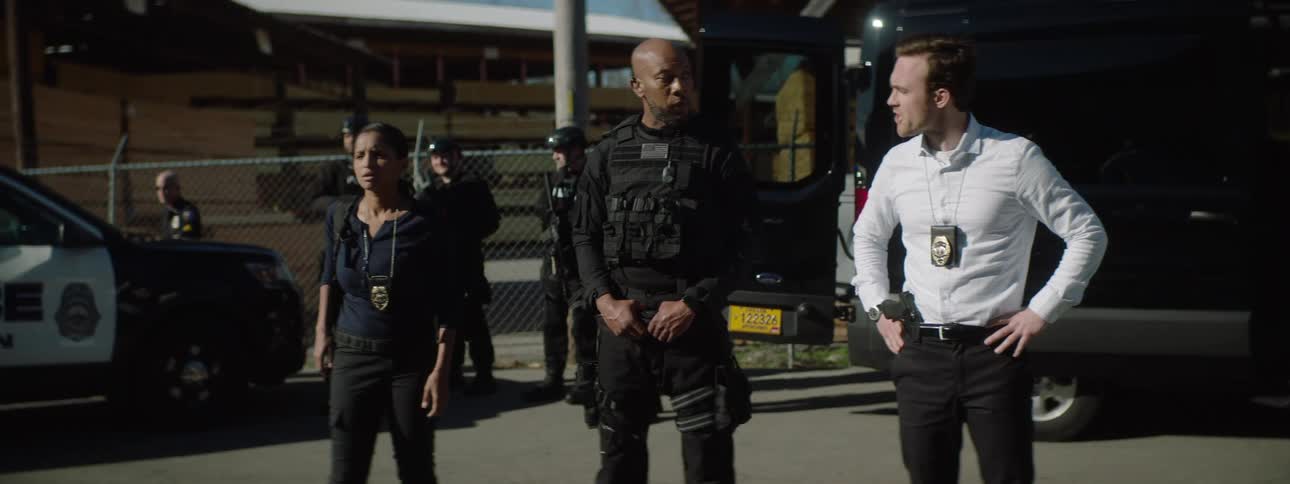 A group of law enforcement officers stands outside a black van with one person in the center speaking while a person on the right has hands on hips