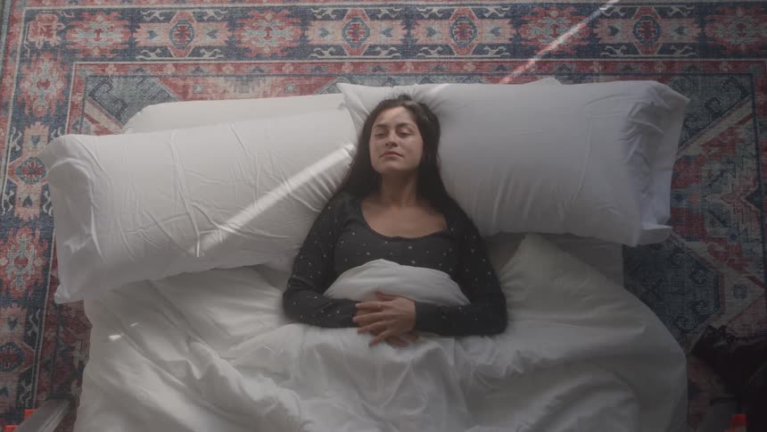 A woman lies down on a mattress her head landing on two fluffy pillows
