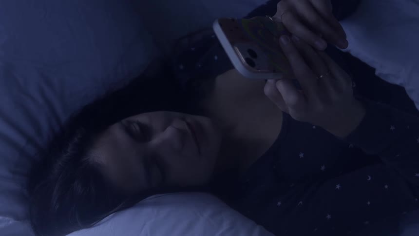 A woman lays in bed in the dark while looking at her cell phone