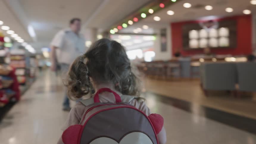 A close rear view shows a young girl with pigtails wearing a backpack with a monkey face walking through a brightly lit airport terminal past stores and people