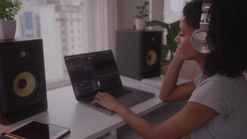 A focused young woman works as a video editor in her home studio wearing professional headphones and typing on a laptop flanked by studio speakers all while sitting at a desk in front of a large city window