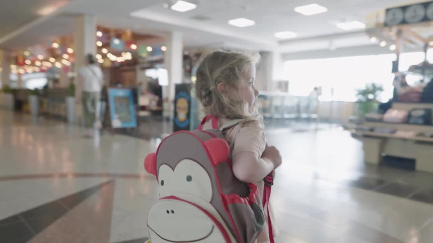 A girl carrying a backpack walks through a corridor in a mall during daytime