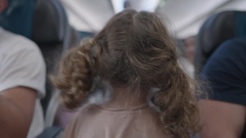 A close rear view shows a young girl with her curly blond hair in pigtails sitting in a plane seat and looking over her shoulder at other passengers