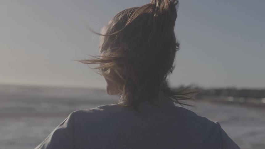 A shot from behind shows a woman with her hair pulled into a ponytail standing on a beach the wind whips her hair around as she looks out at the ocean