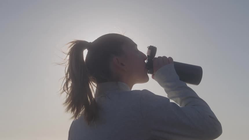Close up of a young woman drinking water from a water bottle while standing outdoors during daytime