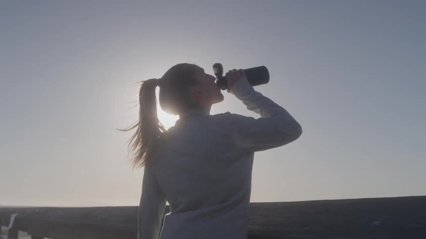 In the video a woman with a ponytail is standing on what appears to be a pier or boardwalk with the sun setting behind her she is seen drinking from a black water bottle