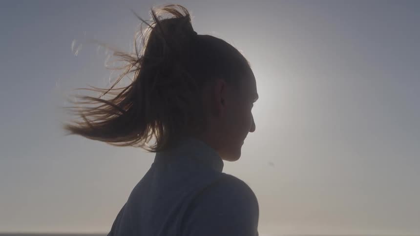 A woman stands in silhouette with her hair blowing in the wind