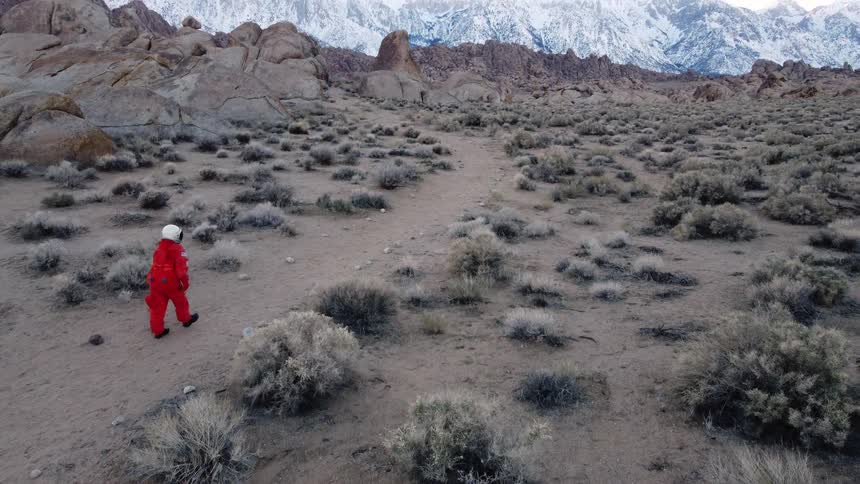 Aerial of an astronaut in an orange spacesuit walking through a wilderness during daytime