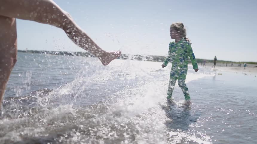 Child playing at the seashore while an adult splashes water with a kick