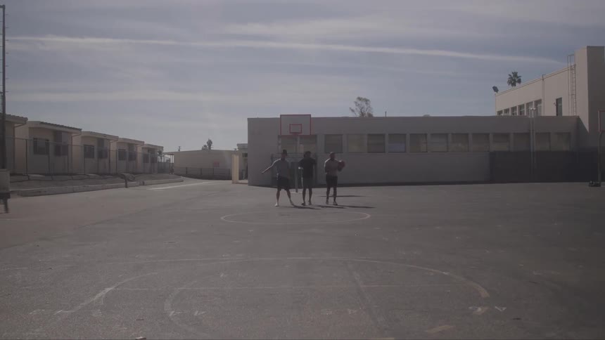 Three young boys are walking on an outdoor basketball court with a basketball hoop on the far end