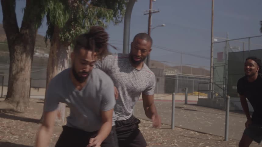 A young man with a beard and dreadlocks dribbles a basketball on an outdoor court while another man attempts to block his shot