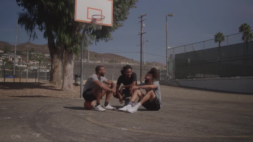 Three young men sitting on a basketball court talking with a ball resting on the ground