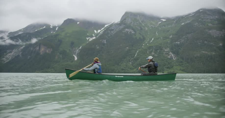 Man and woman paddling on a canoe on a river next to a mountain range outdoors during day time