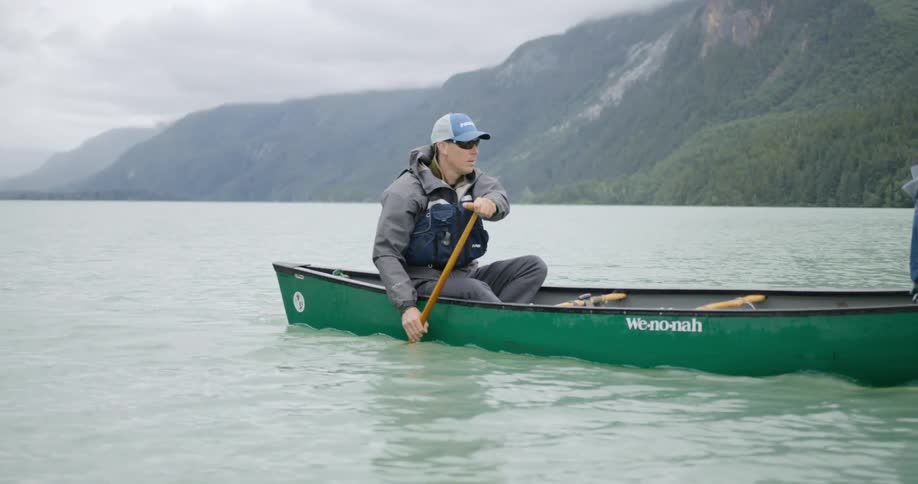 A man wearing a hat sunglasses and a life vest is paddling a green canoe across a large calm lake surrounded by mountains under a cloudy sky