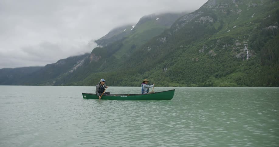 Two people are paddling a canoe across a body of water with large mountains in the background