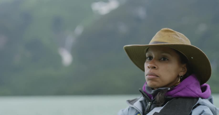 Woman wearing a brown hat and blue jacket casting a fishing line in a lake