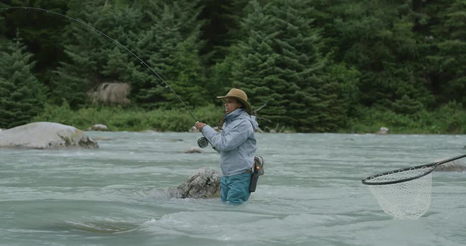 Woman fishing in a river with a fishing rod while someone holds a net nearby