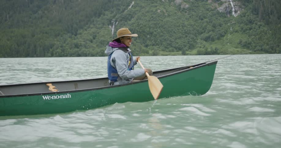 Woman wearing a hat and blue life vest paddling a green canoe on clear waters