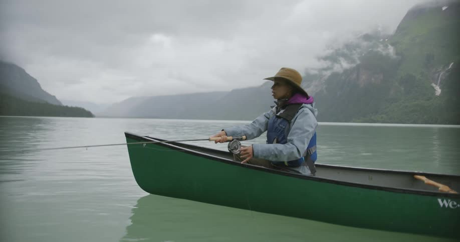 Woman fishing in a green canoe on a lake surrounded by mountains